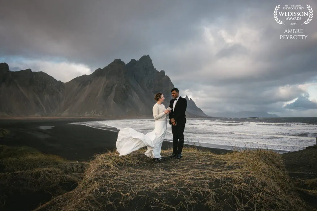 Chic bride and groom exchange vows on a black sand beach with a mountain backdrop, an example of an adventurous elopement