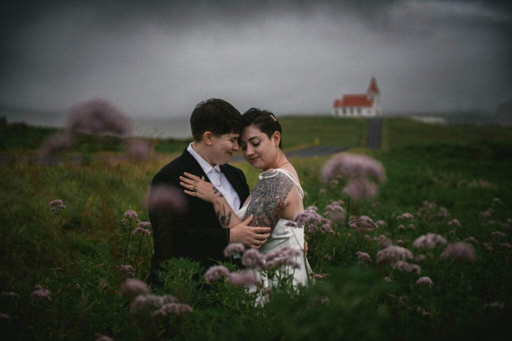 Same-sex couple posing in front of a white and red icelandic church in a field of flowers on a rainy day, on their elopement day in Iceland