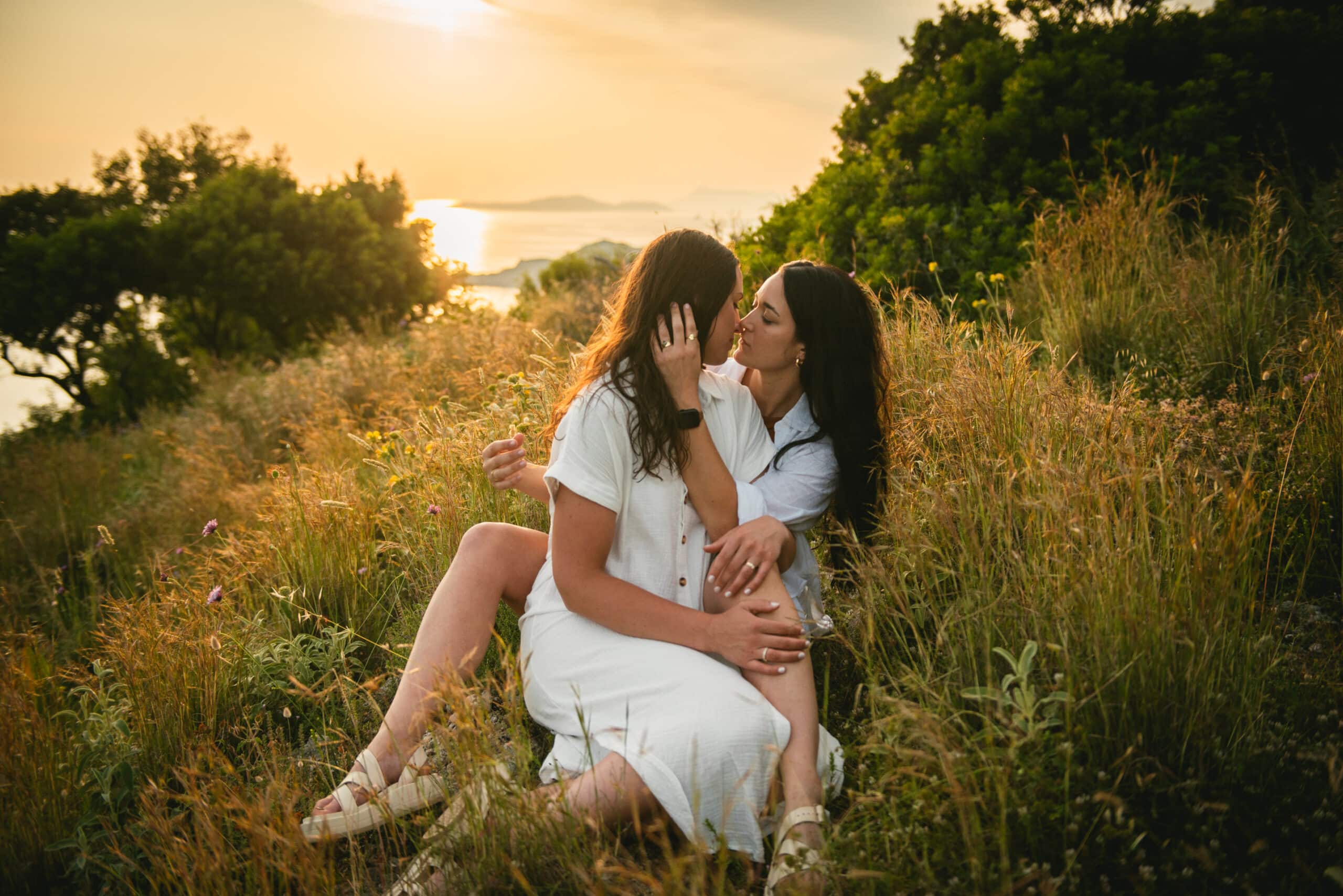 Brides sharing a passionate kiss in the lush grass, capturing their love in Corfu's natural beauty.