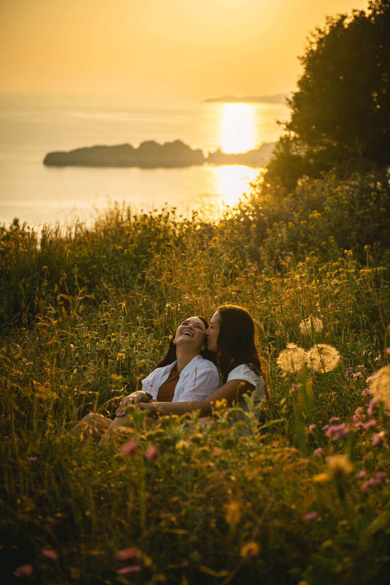 Brides sharing a tender hug while seated in the soft grass, creating a heartwarming moment in Corfu.