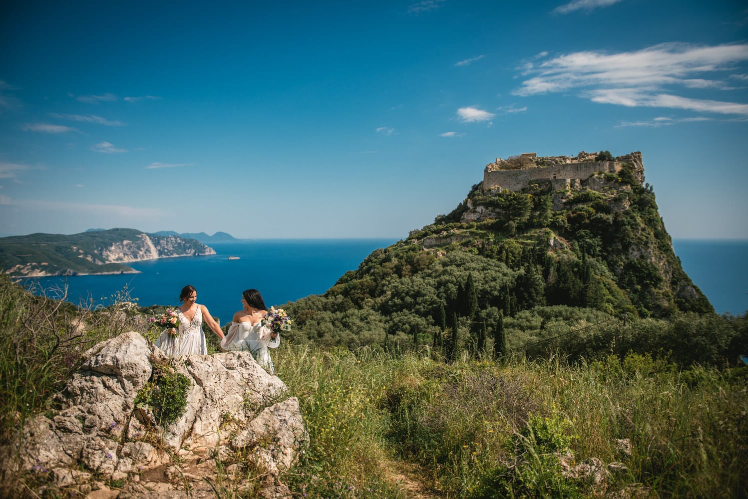 Brides strolling on rocky terrain by the castle, a beautiful Corfu elopement backdrop.