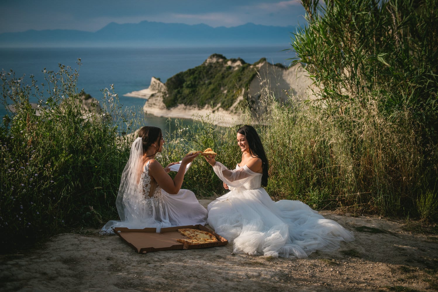 Seaside pizza picnic: Brides share laughter and a meal on the Corfu cliffs during their elopement.