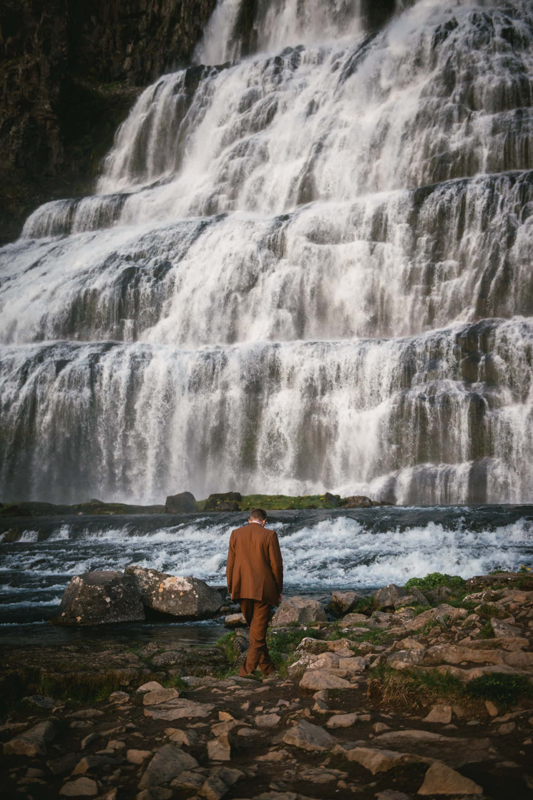 A path towards their vows unfolds, as the groom walks with purpose to the waterfall's embrace, a serene scene within their Iceland Westfjords elopement.