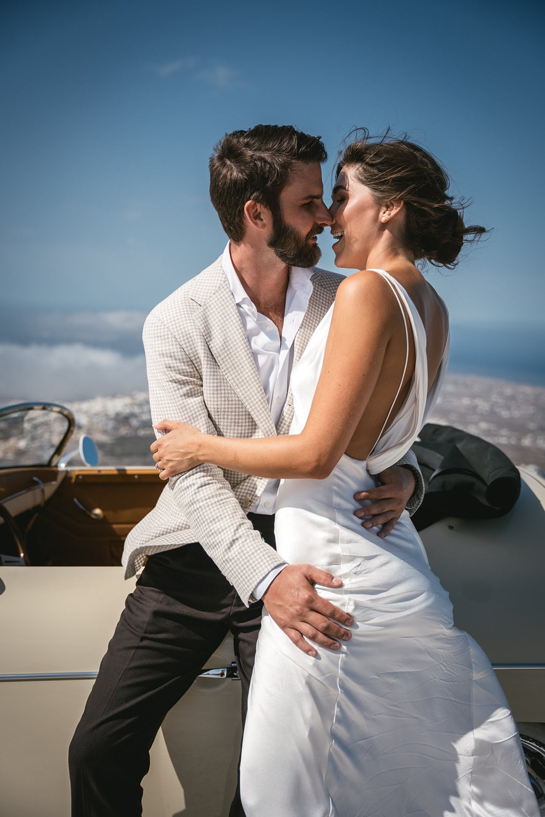 With the island at their feet, they share a kiss, cocooned in the vintage car’s allure.
