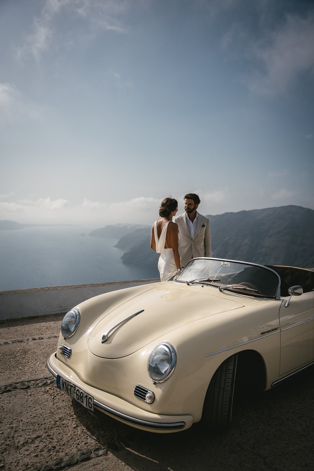 With the azure sea behind, their love story takes a picturesque turn beside the vintage car