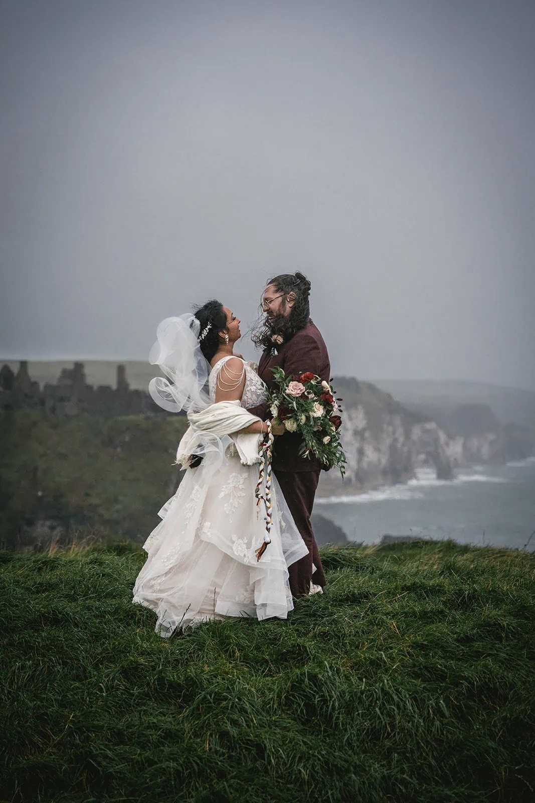 Bride and groom standing in a rain-soaked meadow near Dunluce Castle during elopement
