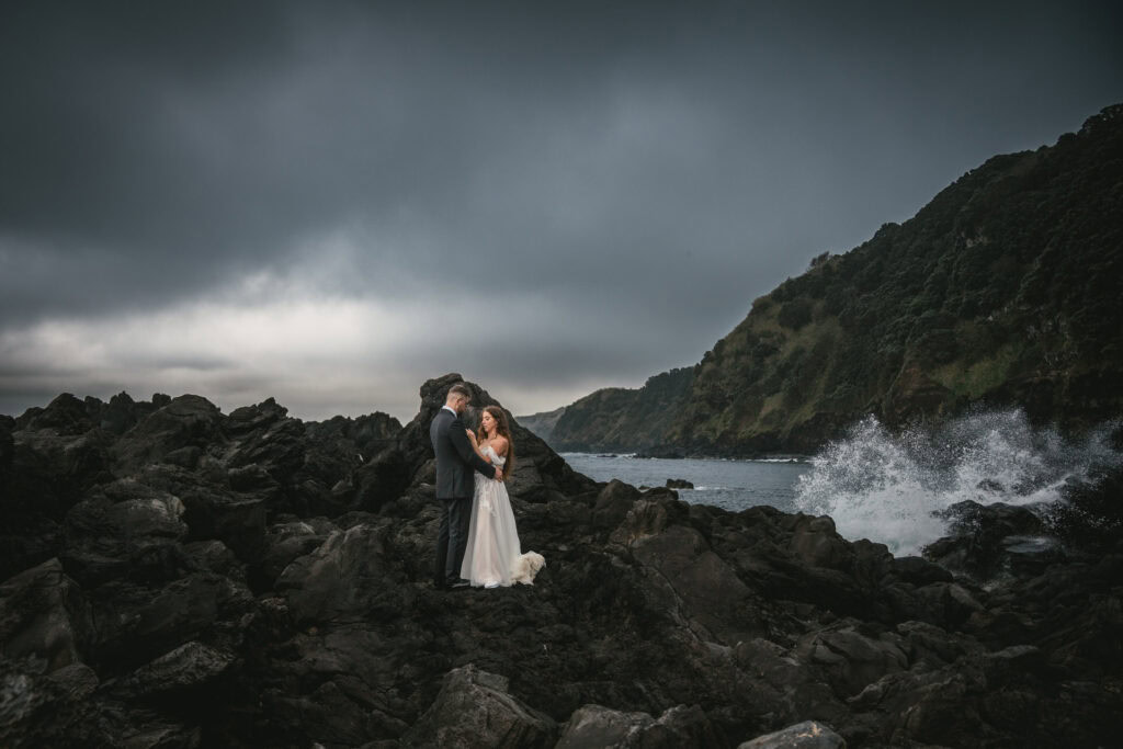 Romantic wedding photo of a bride and groom on rugged rocks by the ocean under moody skies.
