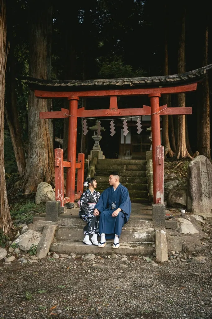 Groom and bride in traditional attire at a serene torii gate surrounded by tall trees.