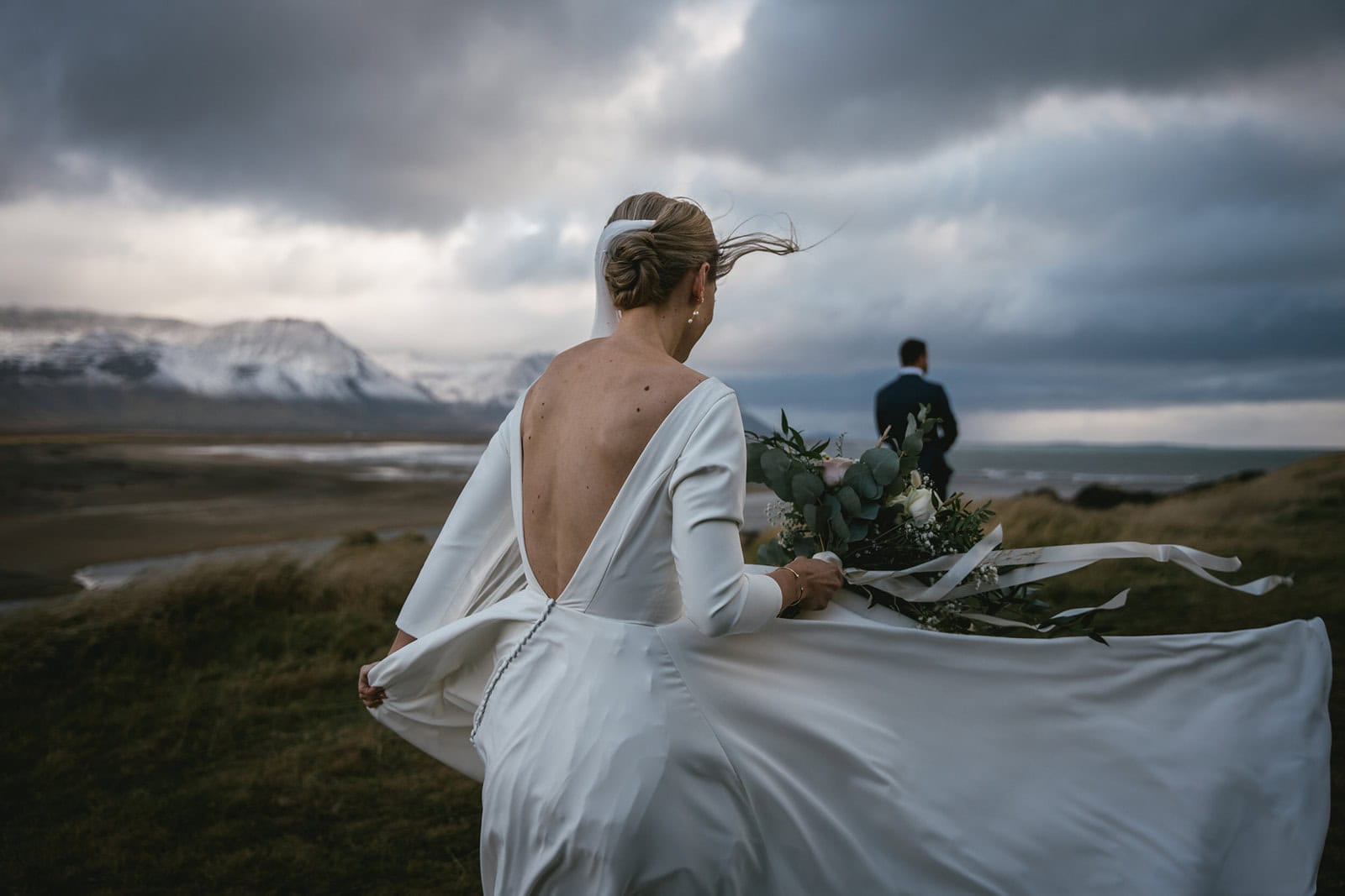 Bride in a flowing white wedding dress with an open back, holding a bouquet, overlooking a dramatic.