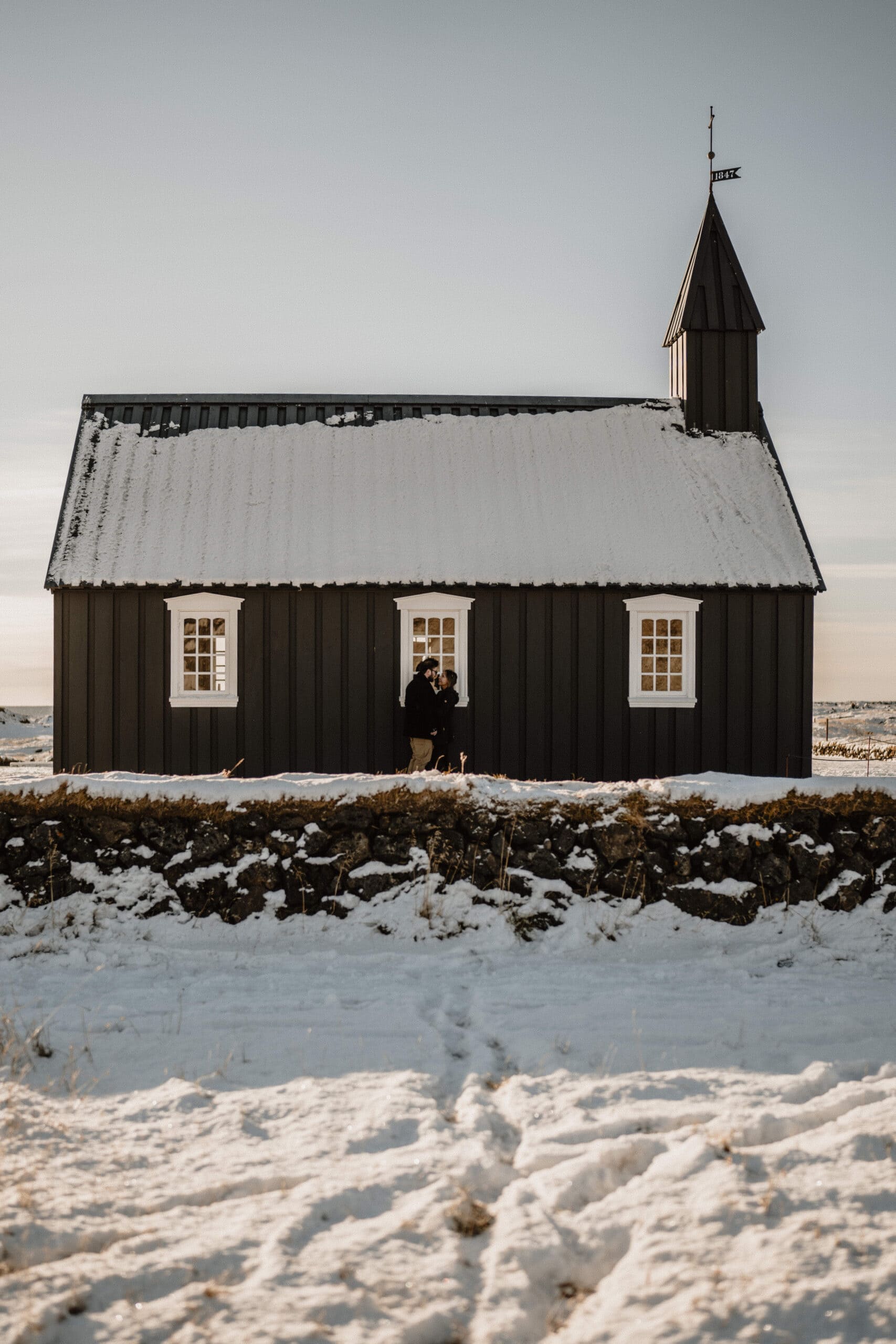Elopement couple standing outside a black chapel in snowy Iceland landscape.
