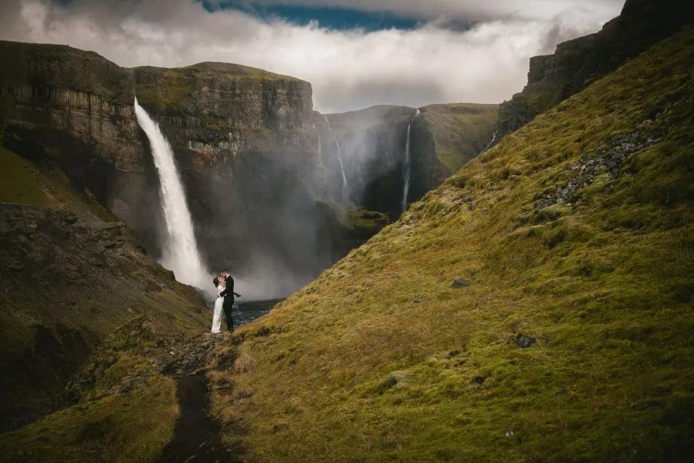 Romantic couple eloping near a stunning Icelandic waterfall with lush green surroundings.