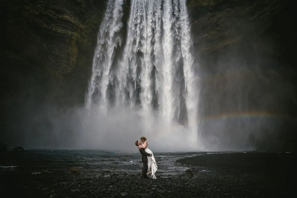 Romantic couple eloping at a stunning Icelandic waterfall with rainbow in the background.