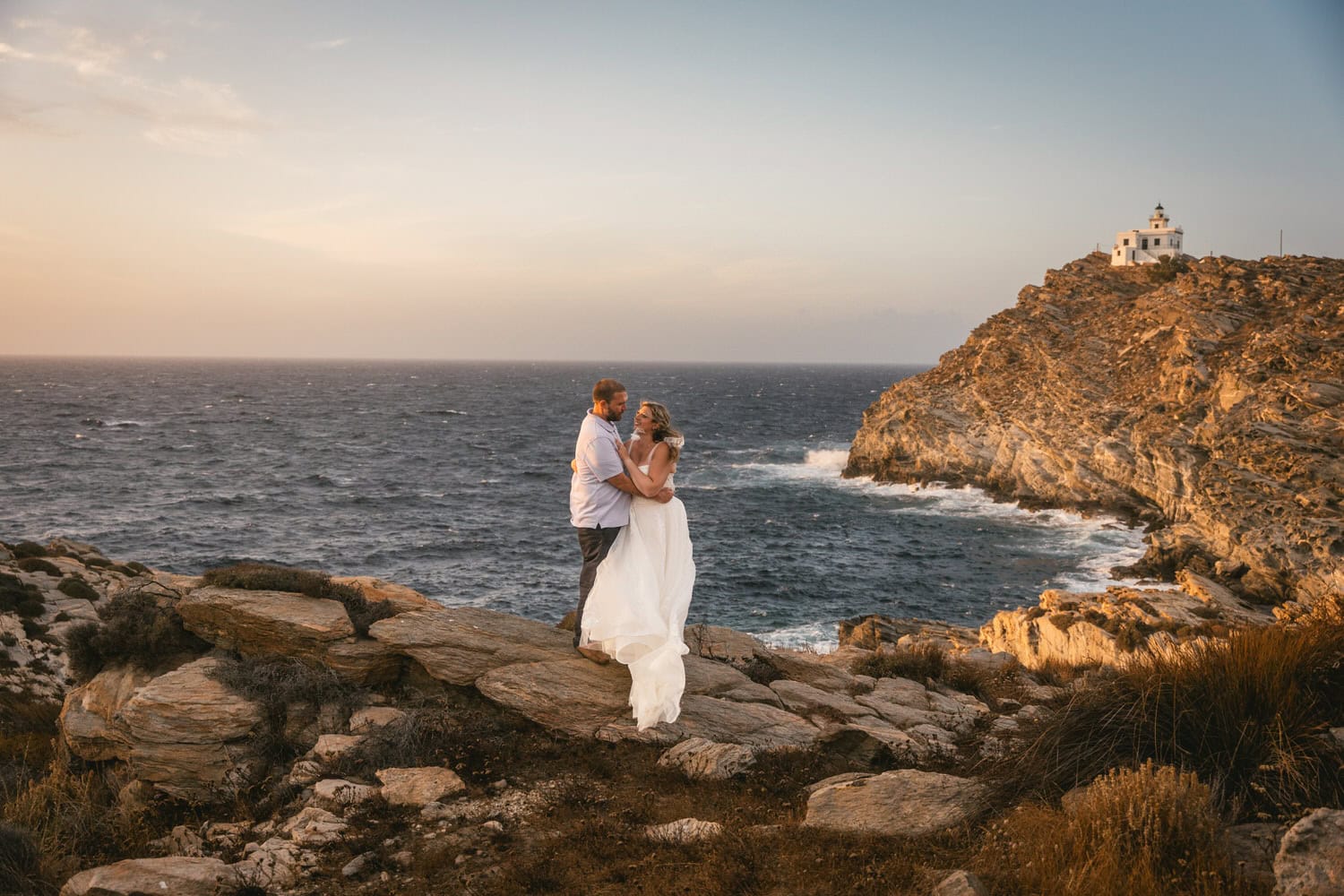 Elegant couple embracing during sunset on rocky coastline in Greece.