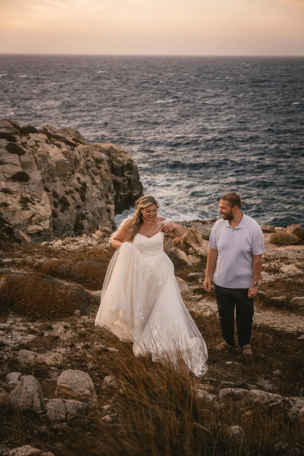 Elopement couple walking along rocky coast at sunset in Greece.