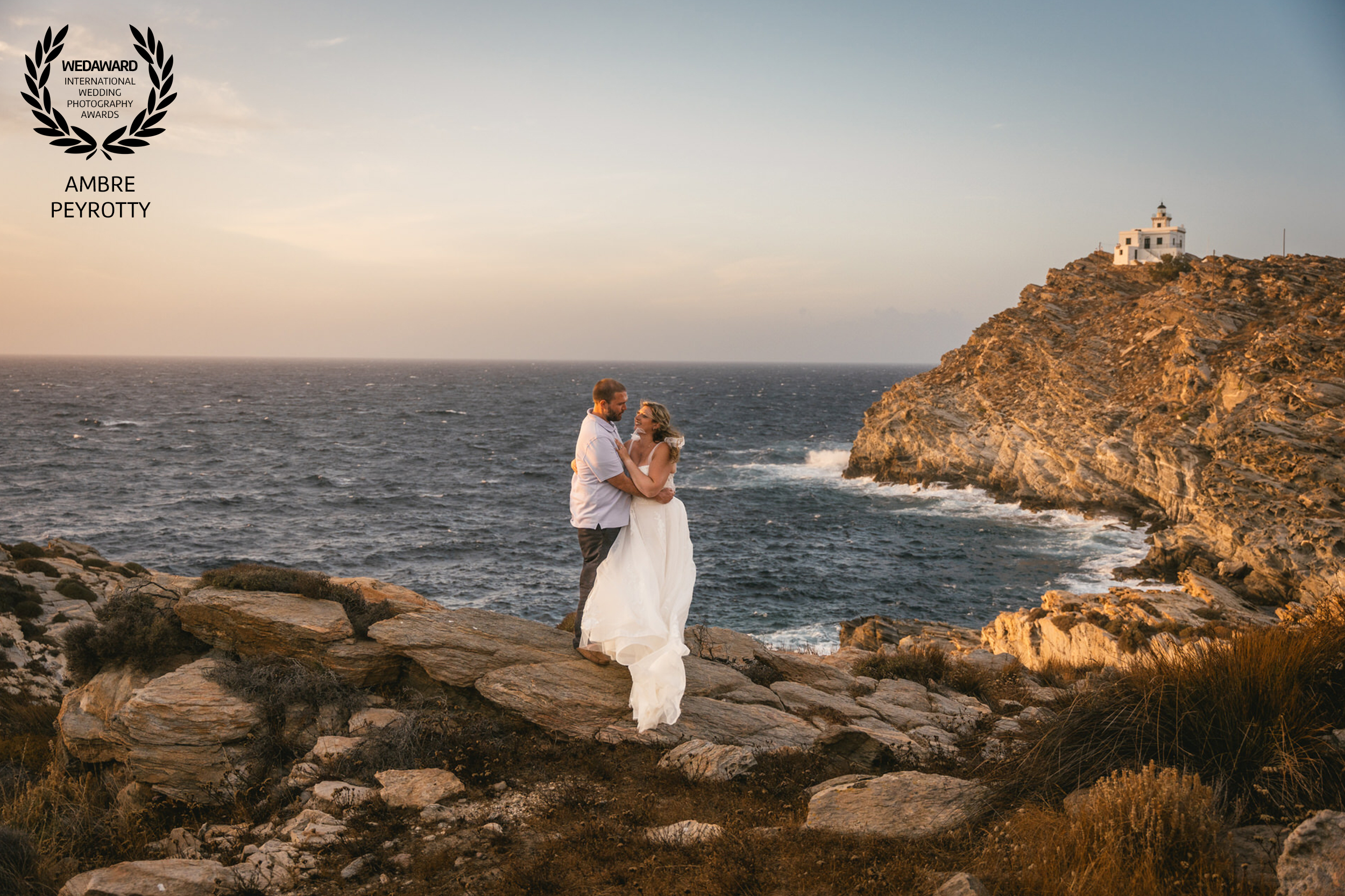 Eloping couple embracing at sunset on rocky coast of Paros, Greece, with lighthouse in background.