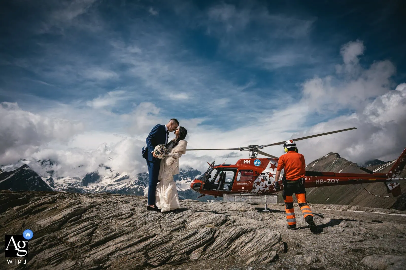 Romantic elopement scene with bride and groom on mountain rock, helicopter rescue team nearby.