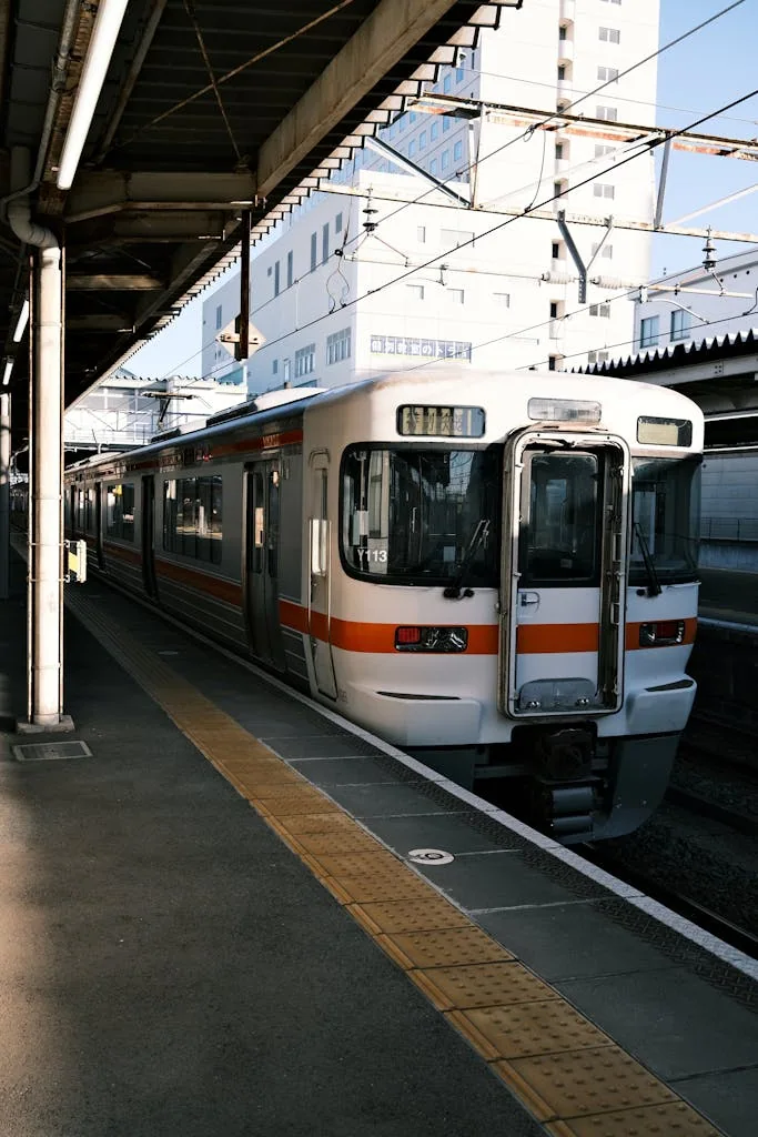 Urban train at city station platform with overhead wires and modern buildings in background.
